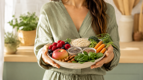 Mujer con ropa verde salvia sosteniendo alimentos ricos en fibra como avena, frutos rojos, manzana, semillas, almendras y verduras frescas en una cocina luminosa de estilo mediterráneo.